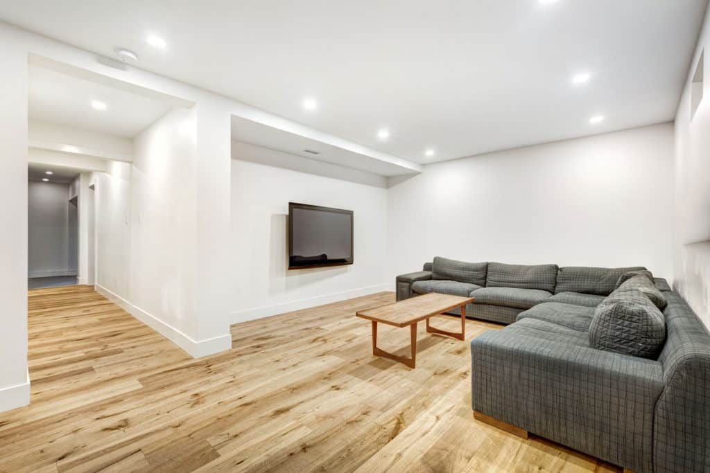 A rustic contemporary inspired basement with wooden flooring, long gray sectional sofa, and a mounted TV