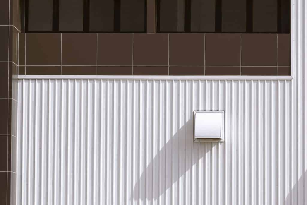 Dryer vent on white corrugated metal wall with row of glass windows