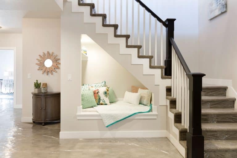 A green carpeted basement stair with white painted wall with a small resting area underneath the stairs, How Wide Do Basement Stairs Need To Be?