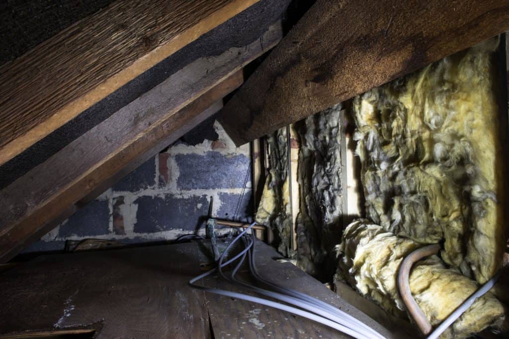 Crawl space under the eves of a house showing old fibreglass insulation, pipework, rafters, breezeblock construction and old boarding.