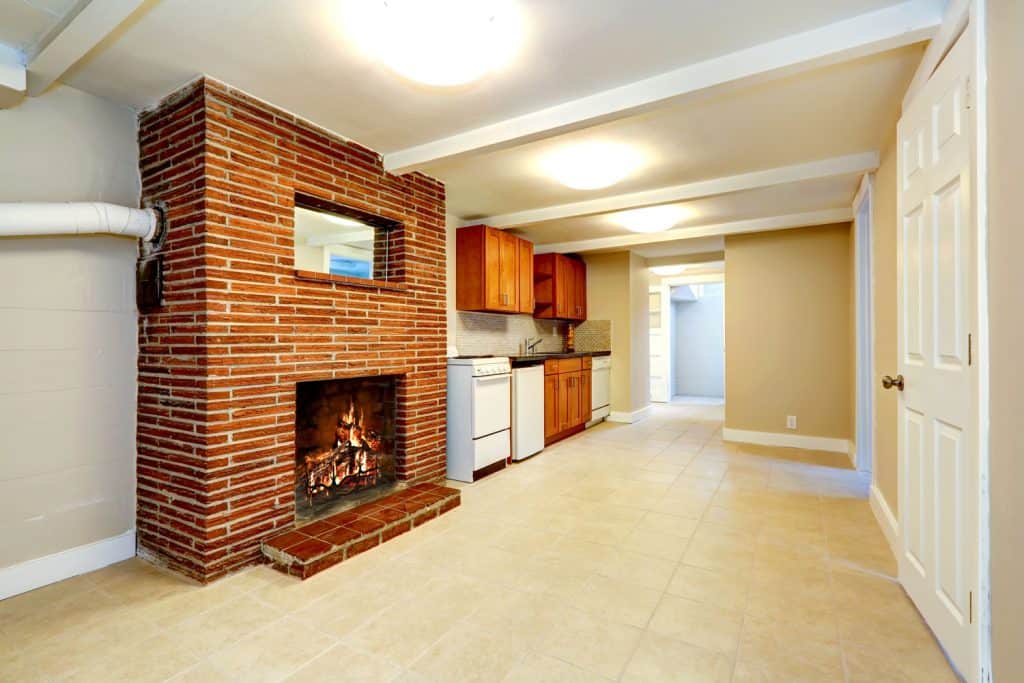 Empty basement room in soft ivory color with tile floor, kitchen cabinets and brick fireplace
