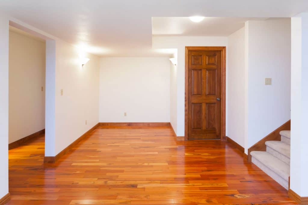 Interior of an empty basement with wooden flooring, white walls, and a hardwood door