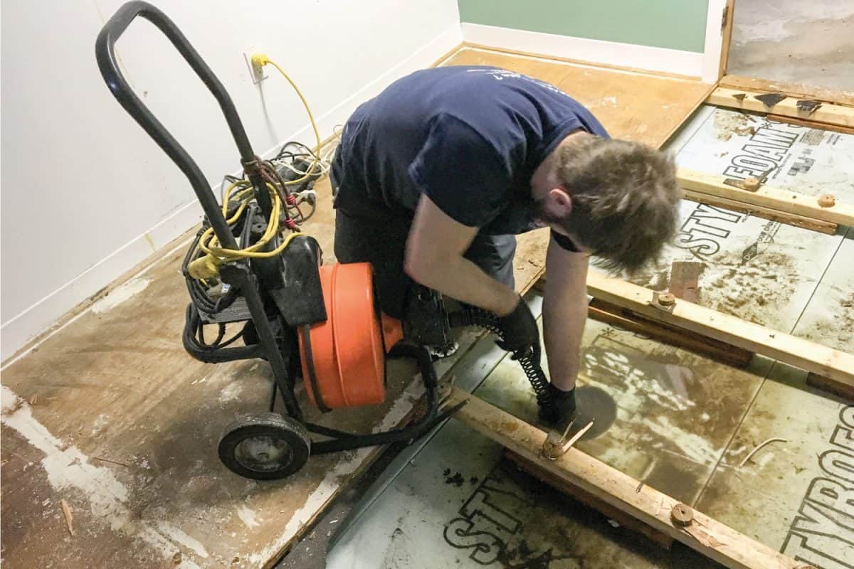 Plumber on his knees in residential home basement and pushing a plunger drain into drain to unclog it