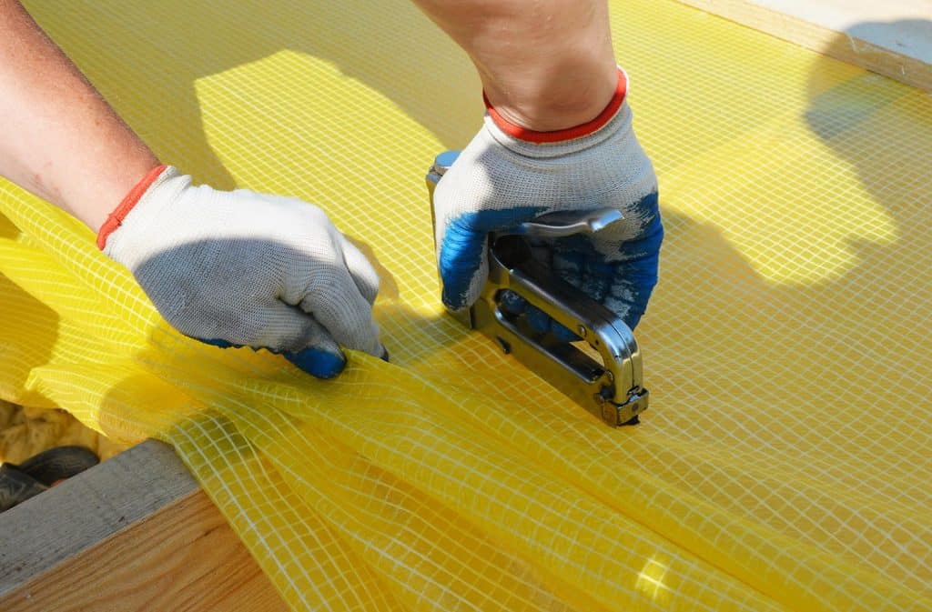 A building contractor is installing vapor barrier, plastic sheeting stapling it to ceiling joists constructing a pitched roof with mineral wool insulation