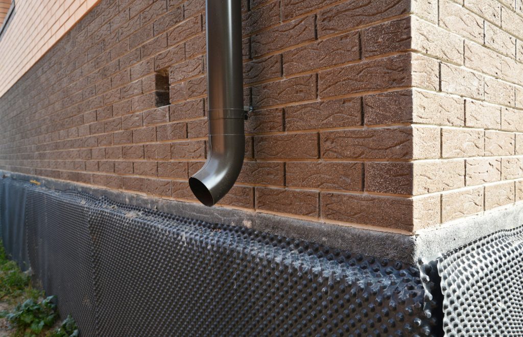  close-up of a metal downspout, roof gutters downspout installed near unfinished foundation, basement covered with a bitumen waterproofing membrane.